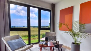 A modern room with a large window, a sofa, a small table with books and a cup, and a potted plant. - Welcome to Backsteinquartier D in Putbus on Rügen (Putbus)