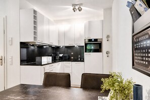 A modern kitchen with white cabinetry, a black countertop, and a built-in oven. There is a dining table with two chairs and a vase with greenery.