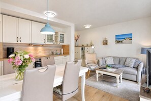 Una cocina moderna con armarios blancos, una mesa de comedor con sillas, un sofá y una mesa auxiliar. Hay un cuadro en la pared y un jarrón con flores sobre la mesa.