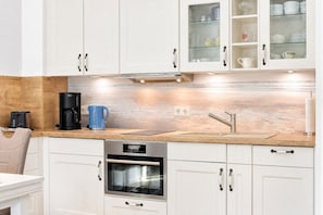Eine moderne Küche mit weißen Schränken, einer Holzrückwand, einem Einbauofen, einer Kaffeemaschine und einem Spülbecken mit Armatur.