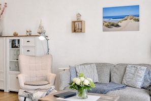 Ein Wohnzimmer mit einem weißen Sessel, einem gemusterten Sofa, einem kleinen Tisch mit Blumen in einer Vase, einem Bücherregal und einem gerahmten Bild von einem Strand.