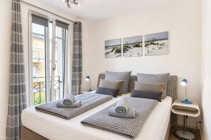 Un dormitorio con una cama, dos mesitas de noche, una lámpara de pared y un ventanal con cortinas.