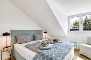 Un dormitorio con techo inclinado, cama con colcha azul estampada, dos lámparas de noche y vistas a árboles a través de una ventana.