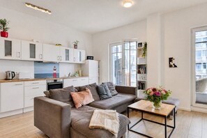 Ein moderner Wohnraum mit grauem Sofa, einem kleinen Holztisch und einer Küchenzeile mit weißen Schränken und einem Wasserkocher.