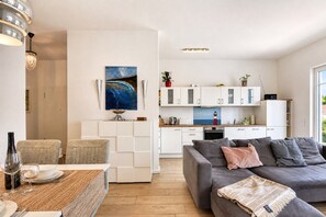 Ett modernt vardagsrum med en grå soffa, en matplats med ett bord för två och ett kök med vita skåp och utsikt över havet.