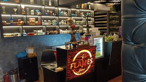 Zona de bar con barra que muestra el letrero 'FLAMES Lounge', estantes con varias botellas y vasos, y un área de asientos con una silla acolchada azul.