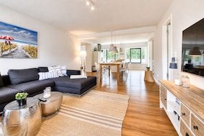 Un salon moderne avec un canapé d’angle, un coin repas et une cuisine visible au fond.