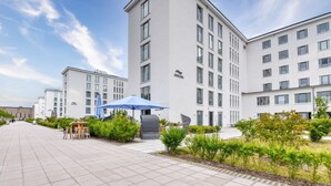 A modern hotel with a clean, white exterior, multiple floors, and a spacious outdoor area with a blue umbrella and seating.