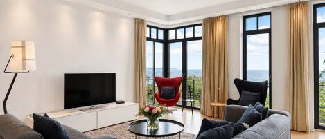 A modern living room with a flat-screen TV, grey sofas, a red chair, and a view of the sea through large windows.