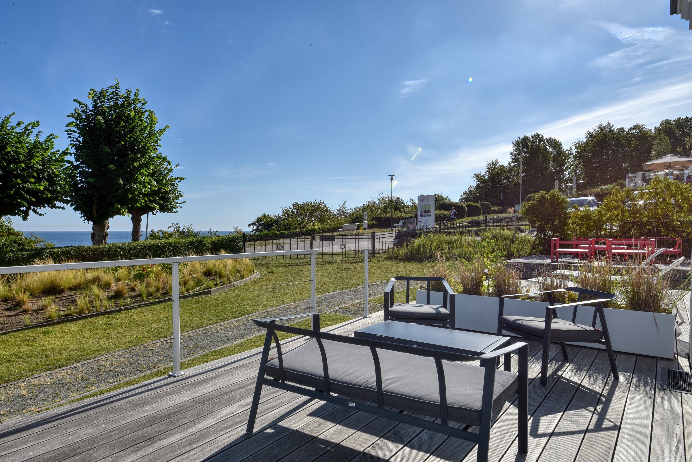 An outdoor seating area with tables and chairs, a wooden deck, and a view of the sea.