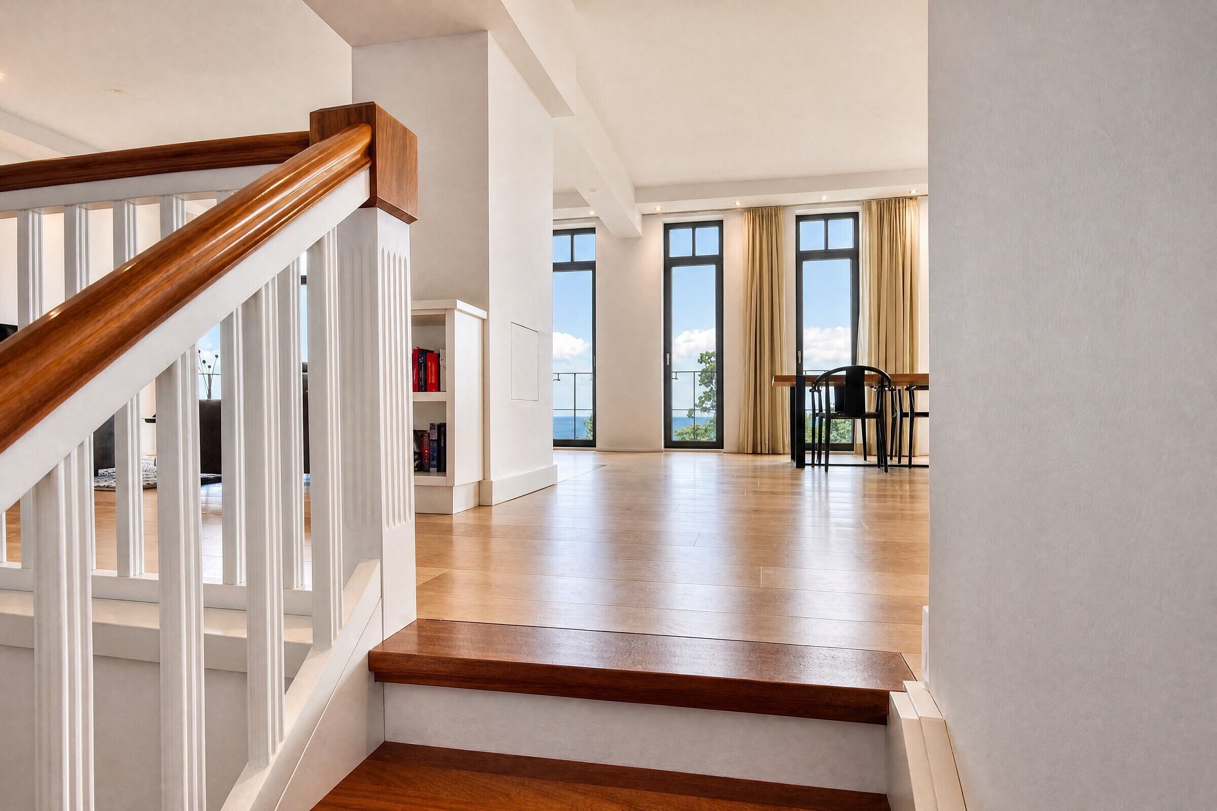 Un amplio salón con escalera de madera, ventanas grandes y vistas al exterior.