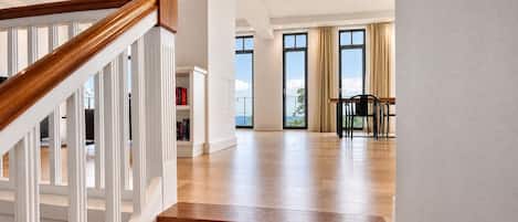 Un amplio salón con escalera de madera, ventanas grandes y vistas al exterior.