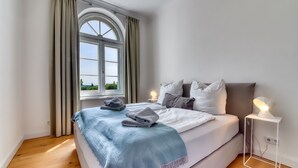 A bedroom with a large window, a bed with a blue blanket, and a bedside table with a lamp.
