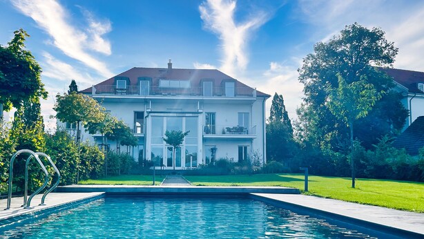 A large house with a swimming pool in front, surrounded by trees and a well-maintained lawn.