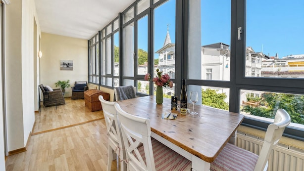 Una habitación amplia con ventanas grandes, una mesa de comedor de madera, sillas blancas, un sofá de mimbre y una planta en maceta.