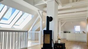 Ein geräumiges Loft mit einem schwarzen Kaminofen, Holzdielen und einer weißen Küche im Hintergrund.