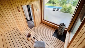 Ein Zimmer aus Holz mit einem großen Fenster, einer hölzernen Bank, einem Eimer und einer Decke mit Muster.