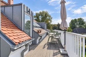 Una terraza techada al aire libre con una mesa y sillas, una pérgola con sombrilla y vistas a árboles y edificios.