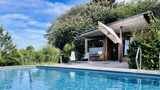 Ein Schwimmbecken mit Holzterrasse, Liegestühlen und einer überdachten Sitzgelegenheit im Freien.