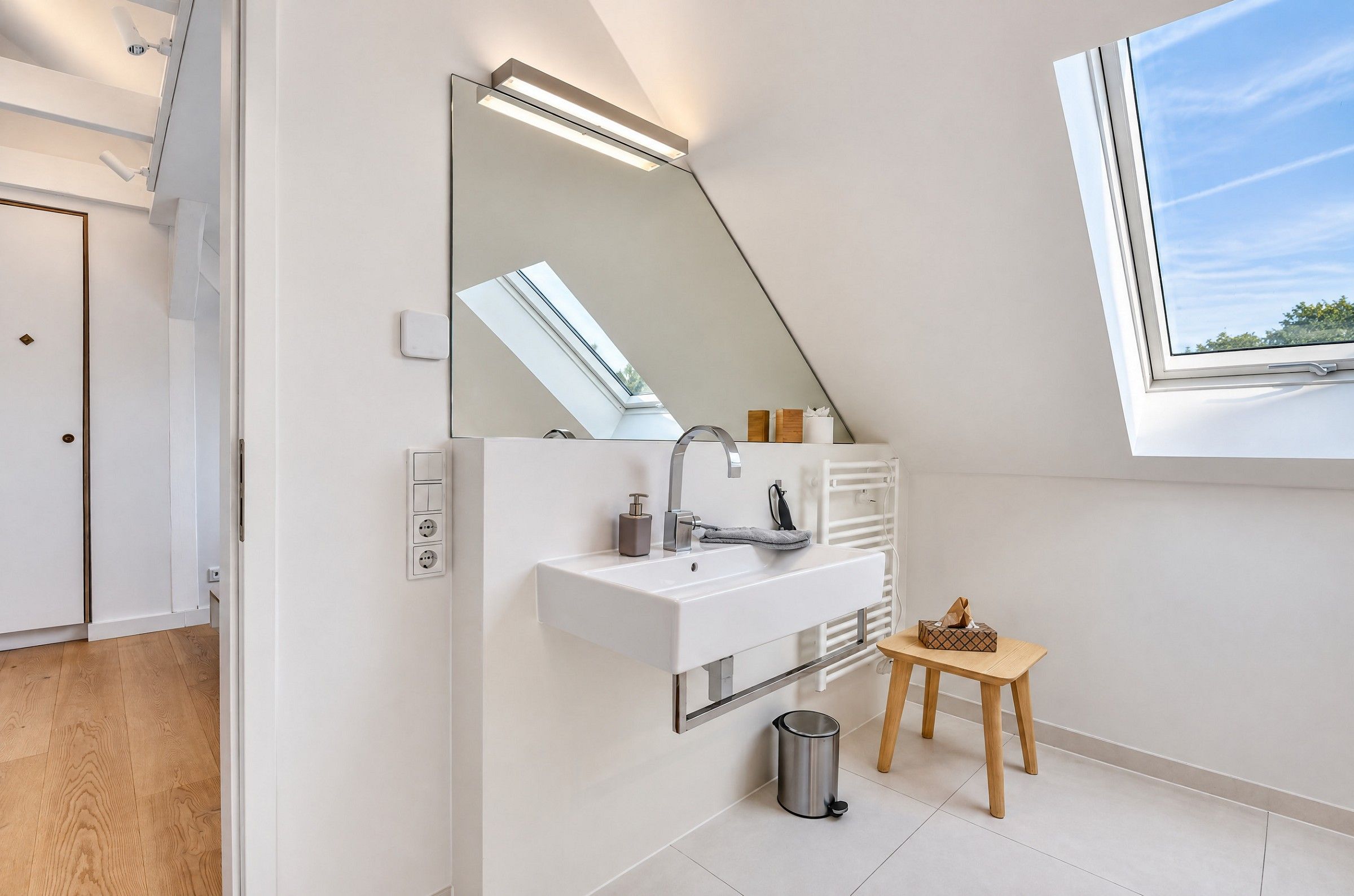 A modern bathroom with a white sink, a mirror, a window, and a small wooden stool.