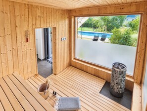 A sauna room with wooden benches, a stone heater, and a view of a swimming pool and greenery outside. - Welcome to our vacation apartment in the heart of Putbus at the Circus! (Putbus)