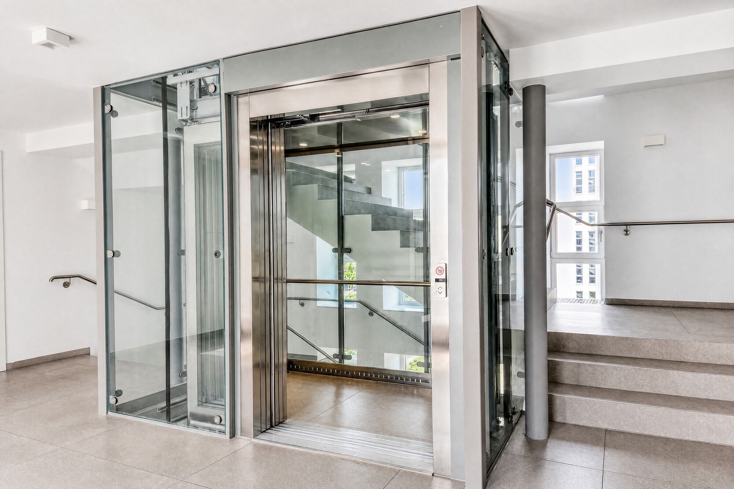A modern elevator with glass doors and a metal frame, leading to a staircase with a metal handrail.