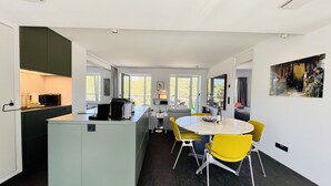 A modern kitchen with a central island, a dining area with yellow chairs, and large windows providing natural light.