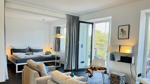 A modern hotel room with a bed, two sofas, a small table, and a view of the outdoors through large windows.