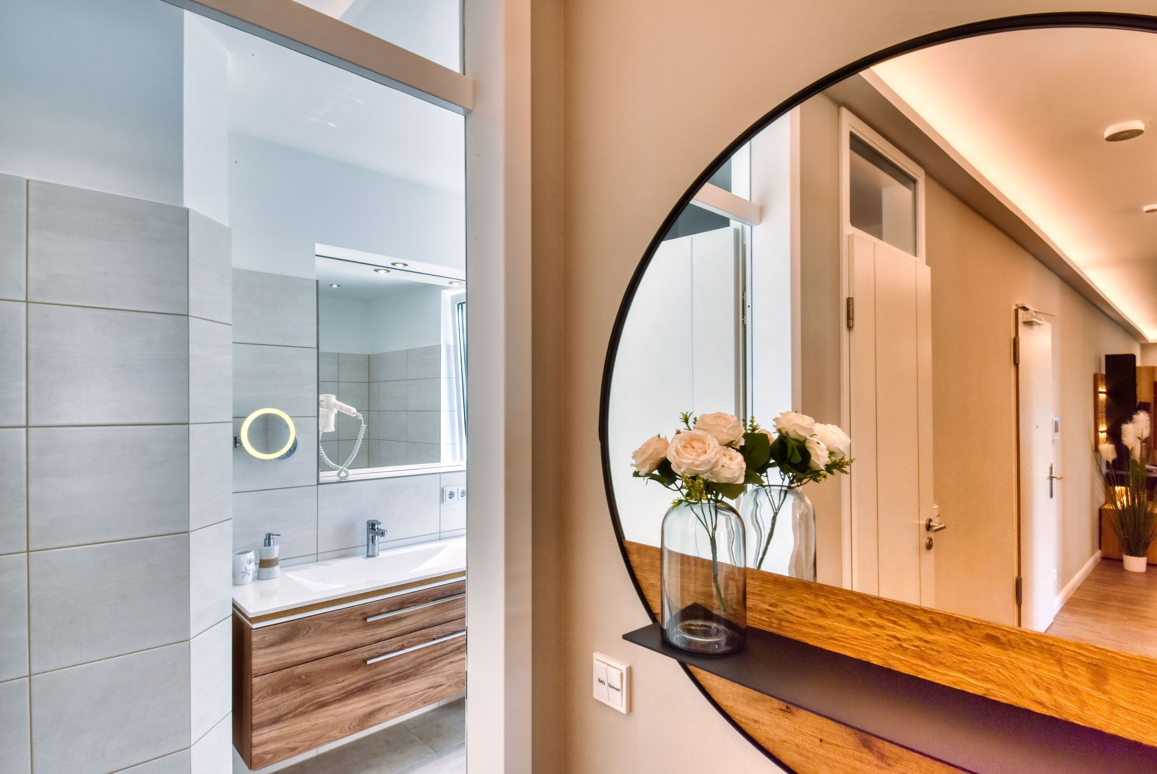 Ein modernes Badezimmer mit einem großen Spiegel, einem Holzwaschtisch und einem Blumenarrangement.