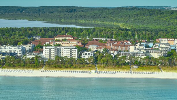 A coastal hotel with multiple buildings, a beach, and a forested hill in the background. - Baltic Sea vacation in Meeresrauschen 2 / Villa Vogue Binz: Sea view and front row (Binz)