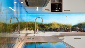 A modern kitchen with a glass backsplash reflecting a beach scene, two stainless steel faucets, and a built-in sink. - Baltic Sea vacation in Meeresrauschen 2 / Villa Vogue Binz: Sea view and front row (Binz)