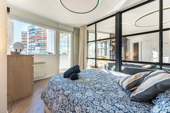 A modern bedroom with a large bed, a wooden dresser, and a view of a cityscape through large windows.