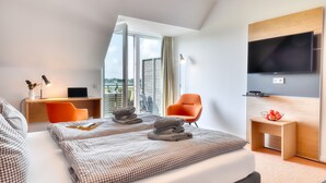 Ein modernes Hotelzimmer mit einem großen Bett, einem Schreibtisch, einem orangefarbenen Sessel, einem Fernseher und Blick auf die Stadt.