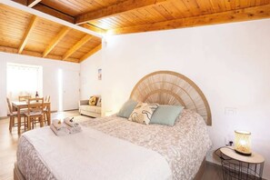 Ein Schlafzimmer mit einem Bett, einem runden Kopfteil, einem Nachttisch mit Lampe, einer Holzbalkendecke und einem Essbereich mit Stühlen und Tisch.