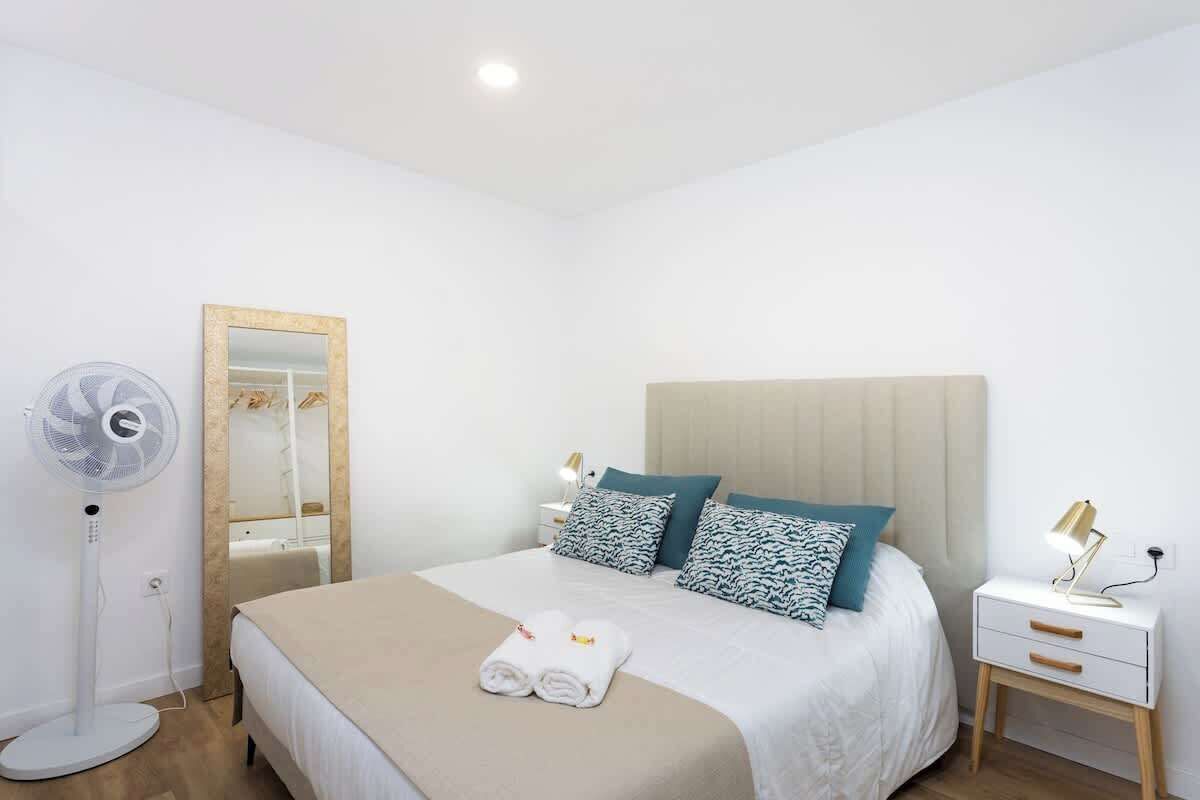 A hotel room with a bed, bedside table, lamp, and a standing fan.