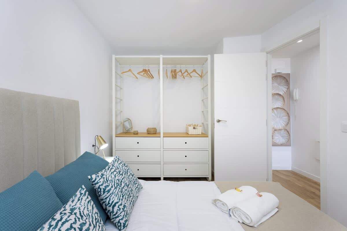 A bedroom with a bed, a bedside table with a lamp, a wardrobe with hanging clothes, and a mirror.