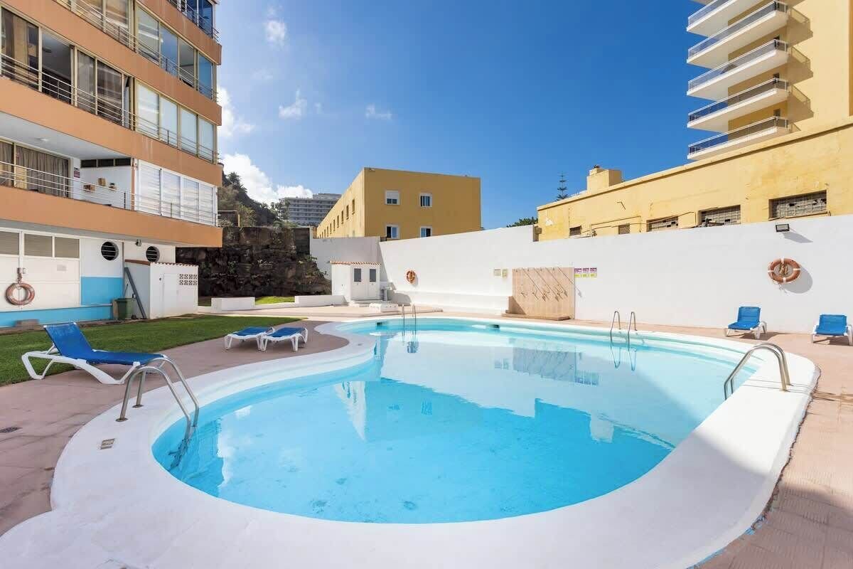 Une piscine avec des chaises longues, un mur blanc orné d’une bouée de sauvetage et, au fond, un immeuble à plusieurs étages.