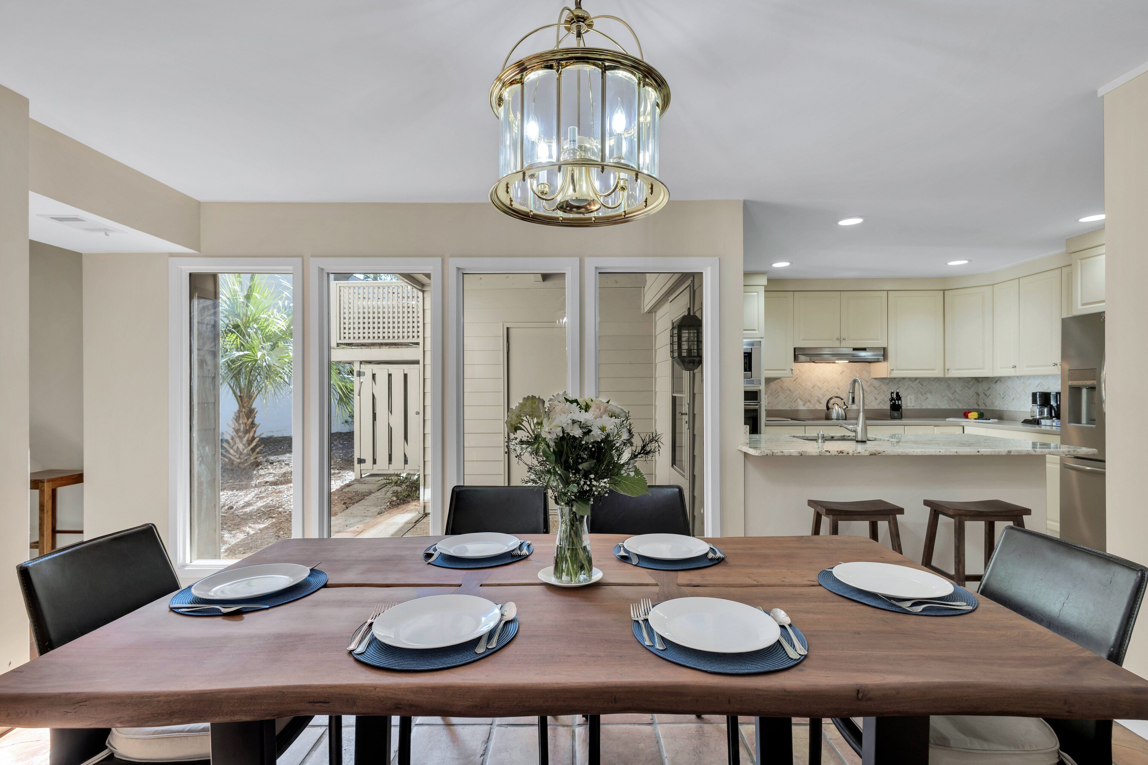 A dining area with a wooden table set for six, a chandelier, and a view of a patio with palm trees.