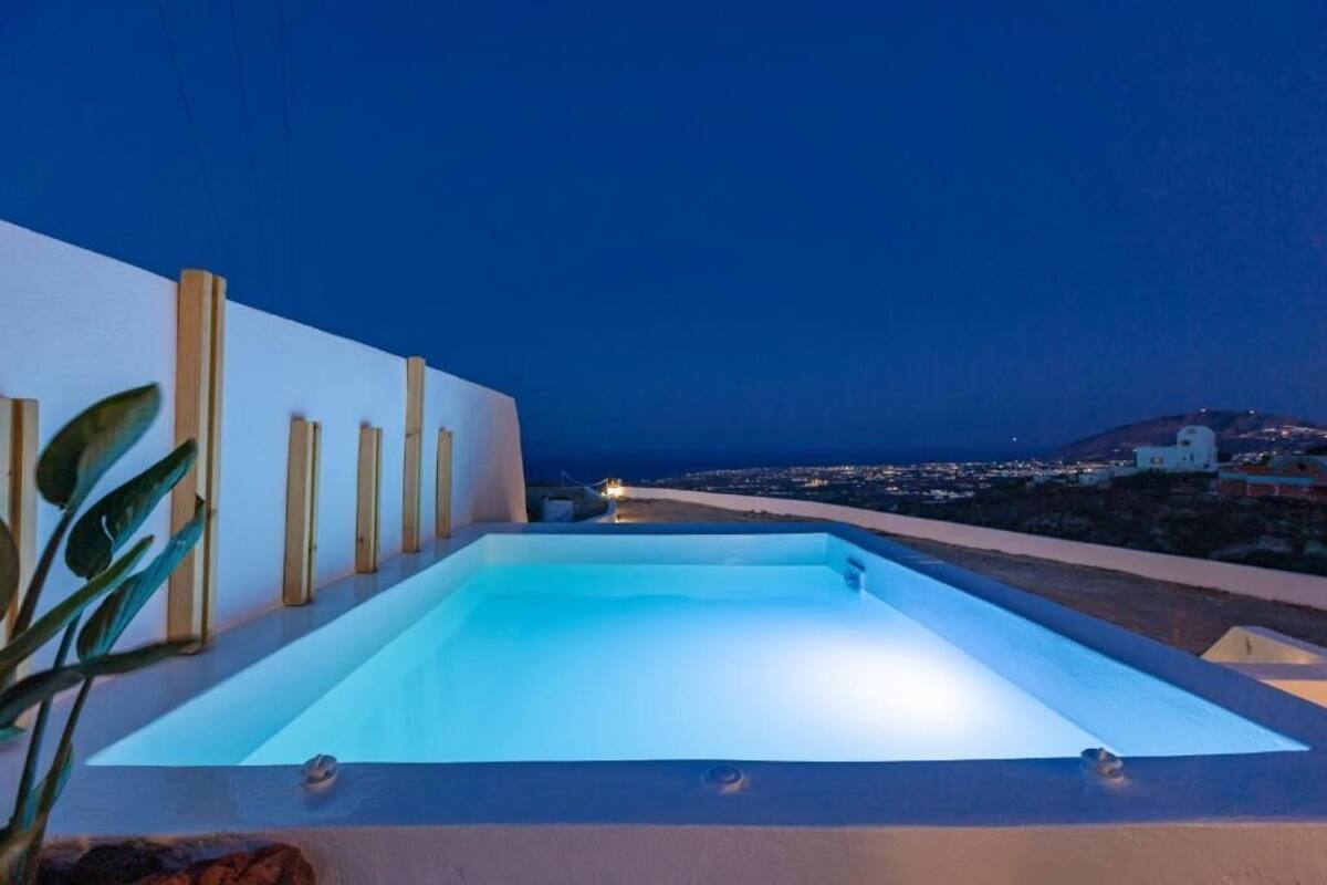 Una piscina rectangular con superficie de agua azul cristalina, rodeada por un muro bajo y postes de madera, con vistas a un paisaje urbano nocturno.
