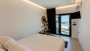 Een moderne hotelkamer met een groot bed, een aan de muur bevestigde televisie, een tafeltje met een plant en uitzicht op de zee via een schuifdeur.