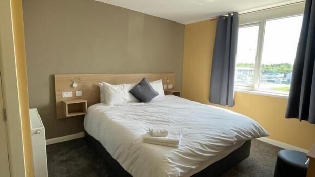 A hotel room with a bed, a bedside table, a lamp, a window with curtains, and a view of the outside.