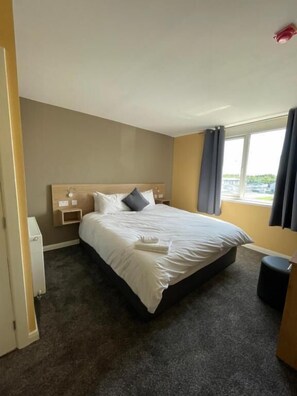 A hotel room with a bed, a bedside table, a lamp, a window with curtains, and a view of the outside. - Tinapa Suites (Manchester)