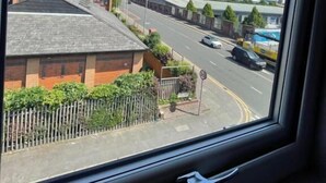 A view from a window of a building with a street in front, including a road, a fence, and some greenery.