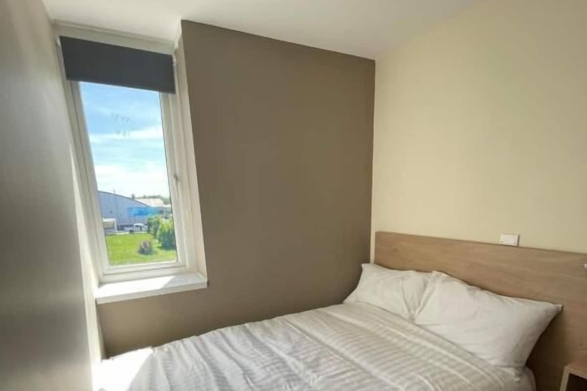 A single bed with a white bedspread and pillows, a small bedside table, and a window with a view of greenery and buildings.