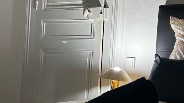 Une chambre d’hôtel avec une porte blanche fermée, un lit recouvert d’une couvre-lit à motifs floraux, un oreiller noir et une lampe de chevet.