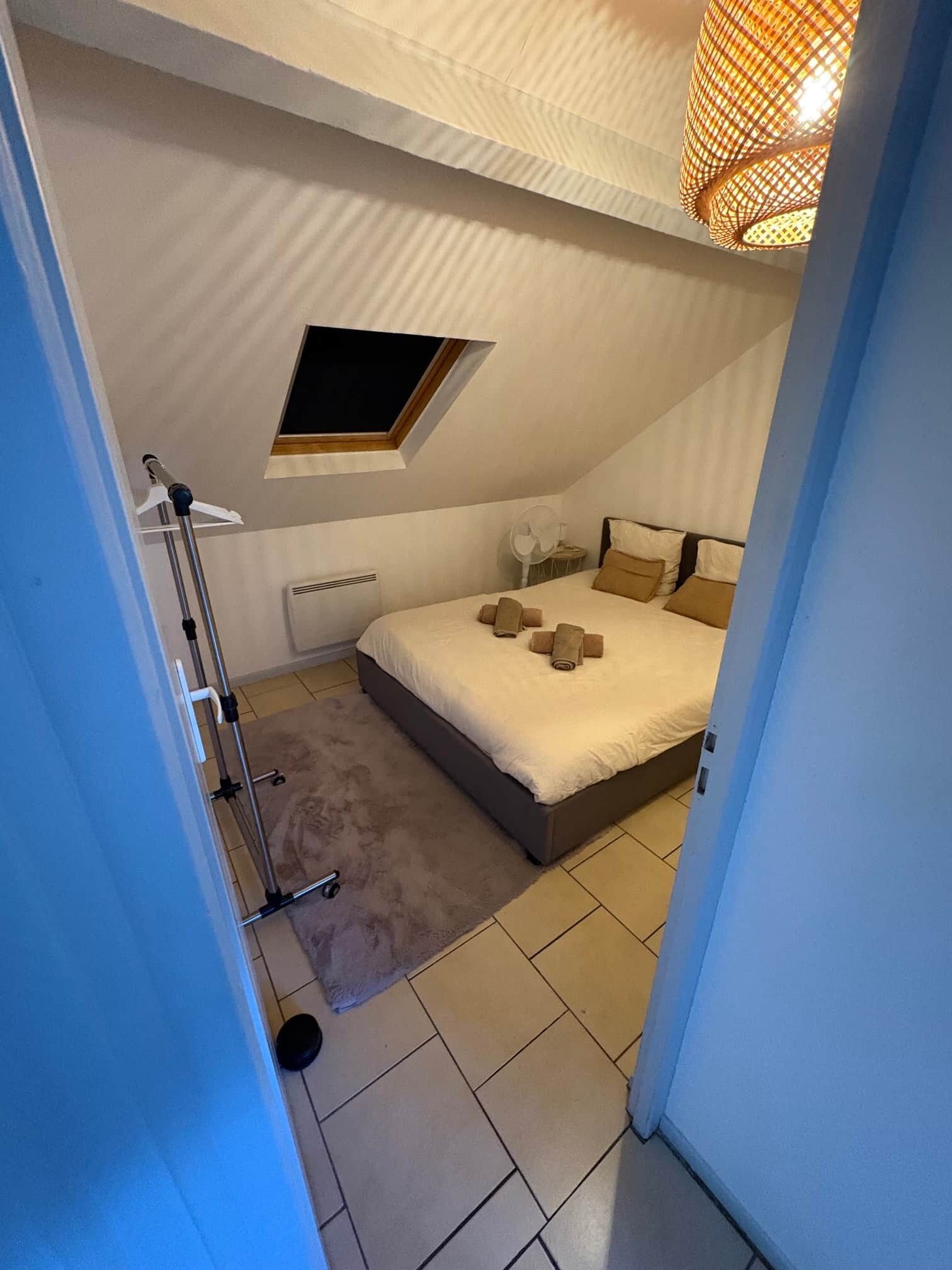 Un dormitorio con una cama, un ventanal pequeño, un radiador y un ventilador.