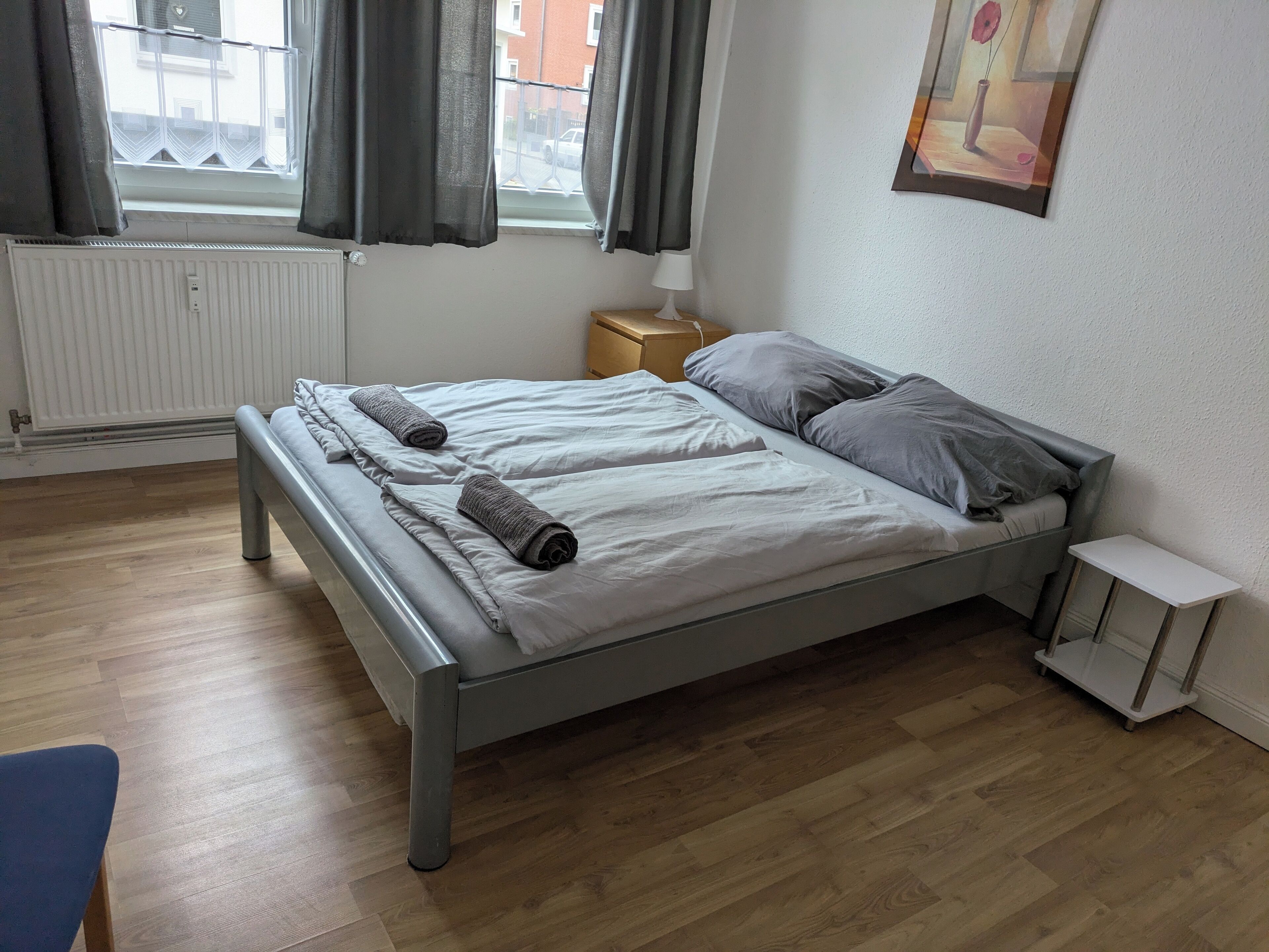 A double bed with grey linens and pillows, a bedside table with a lamp, a radiator, and a window with curtains.