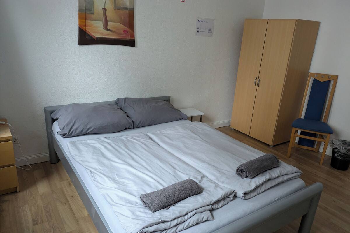 Ein ordentlich bezogenes Bett mit grauer Bettwäsche und Kissen, ein kleiner Nachttisch, ein Holzschrank und ein Stuhl.