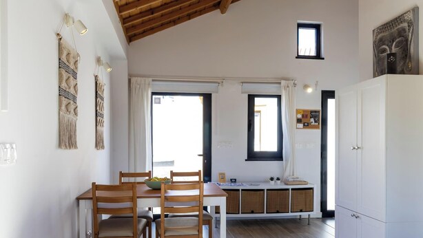 A dining area with wooden chairs, a table, and a bowl of fruit. There are two windows with curtains, a small window above the table, and a cabinet with a picture on top. - Casa da Gente T1 by PontaNegraAzores (Praia da Vitória)