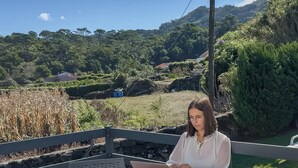 A person using a laptop at an outdoor table with a view of a rural landscape. - Casa da Gente T1 by PontaNegraAzores (Praia da Vitória)